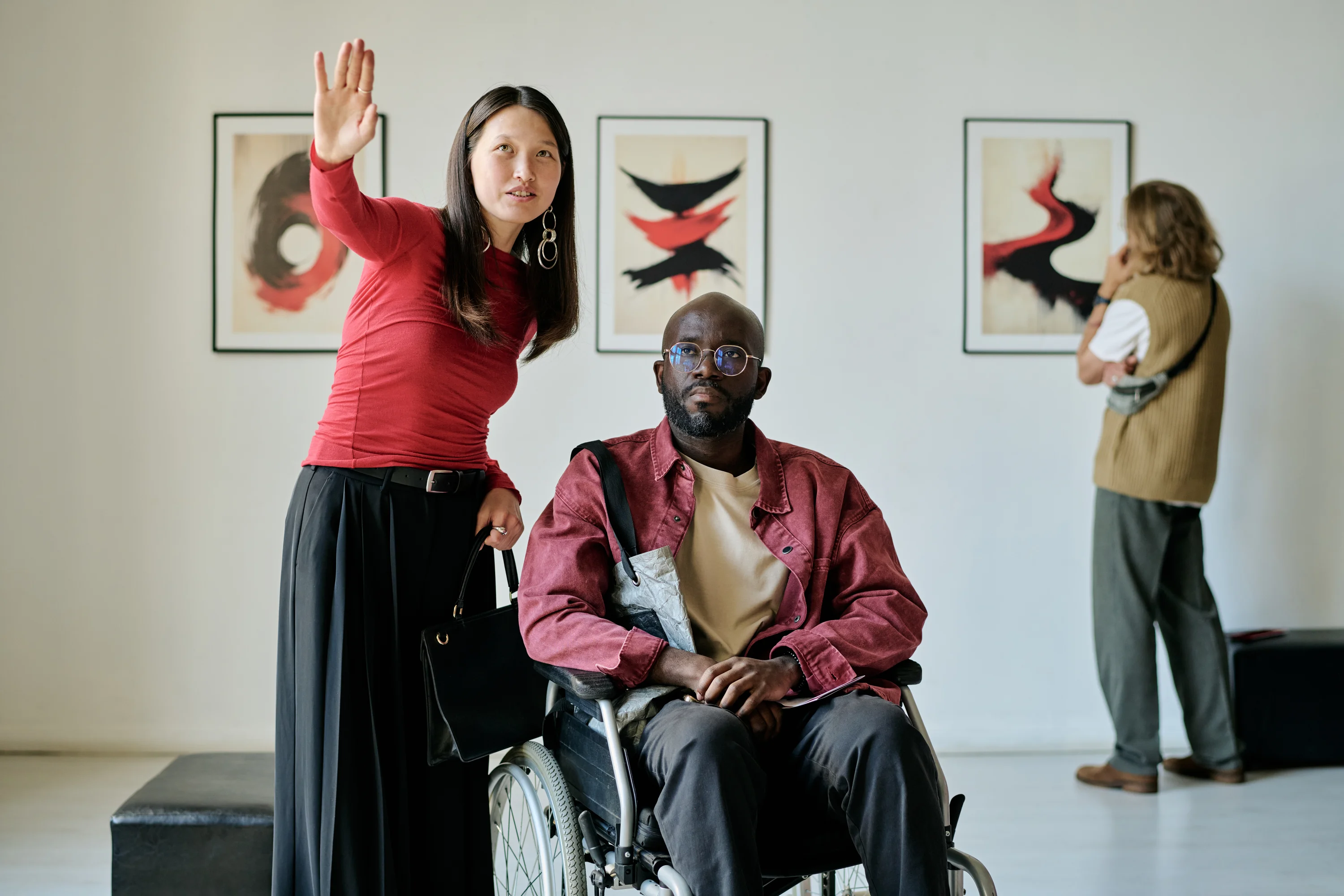 Visitors viewing artwork in a gallery, including a wheelchair user and a companion engaging with the exhibition.