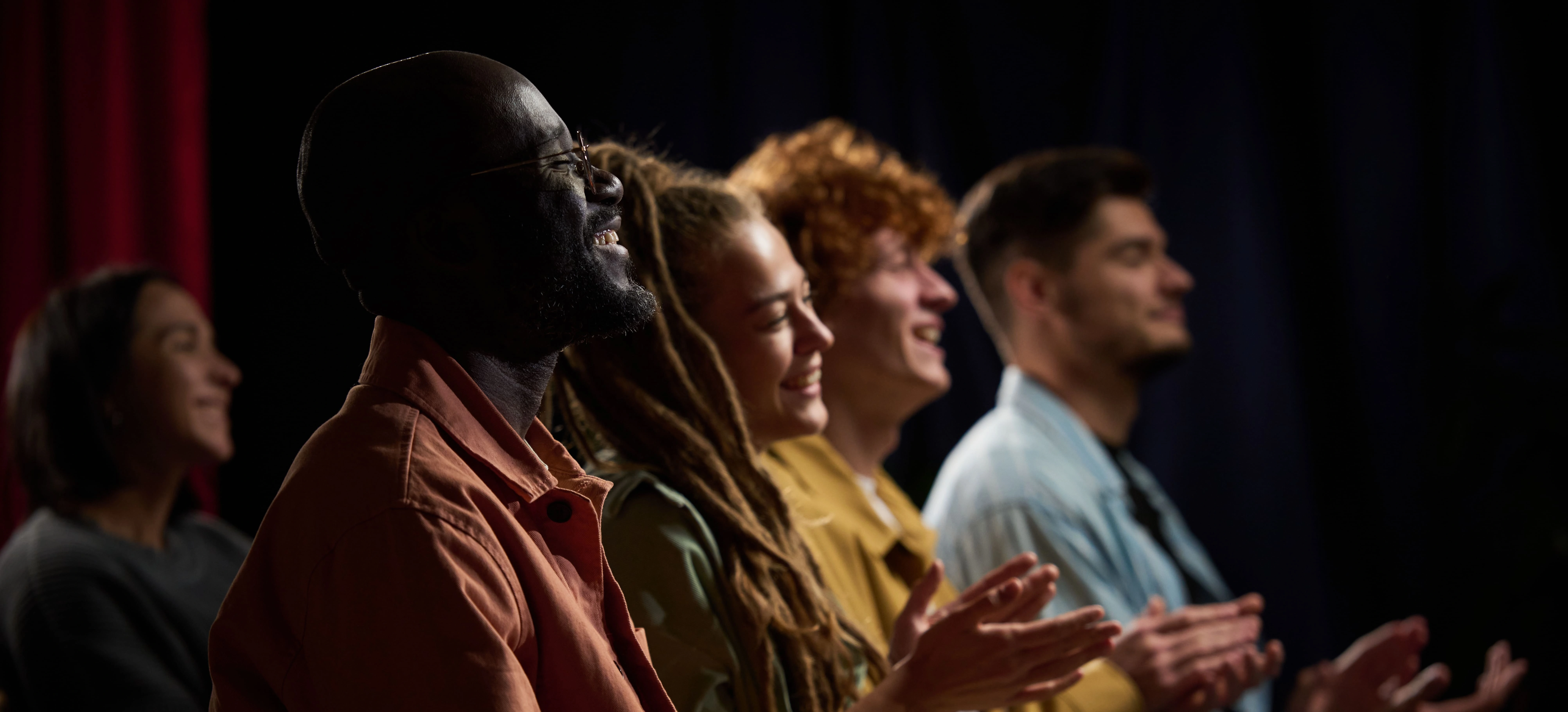 A diverse group of young adults seated in a theatre applaud and smile toward the stage, capturing a positive audience reaction.